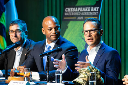 Two men sit at a table. One of them is speaking into a microphone while the other man watches him.