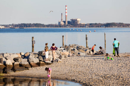 Smokestacks are seen from across a river, where people play along a beach in the foreground.