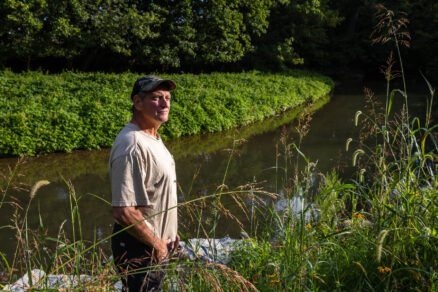 A man stands next to a river in wetland plants. Across the river, green grasses grow.