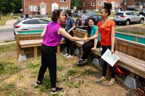 A woman shakes hands with another woman who is holding a map. They are standing in front of a wooden bench which is on grass next to a city street with houses on the other side of the street.