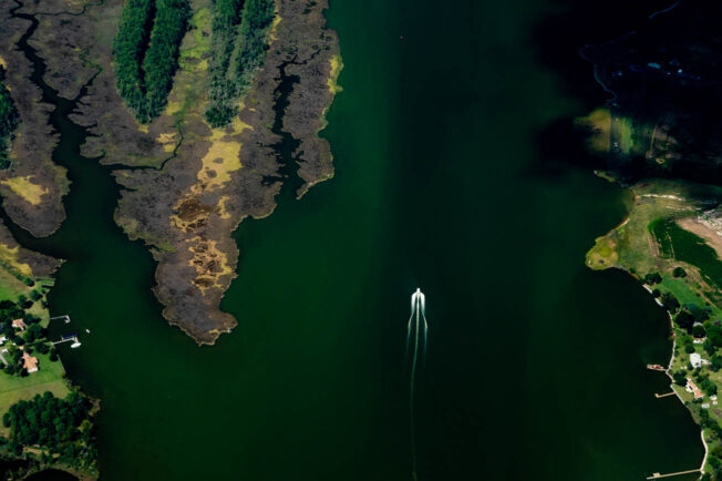 A boat is seen from above, in the middle of a river and avoiding channels near the shore.