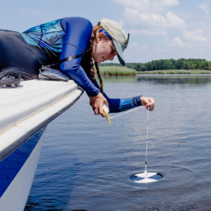 A woman leans over the side of a boat, slowly lowering a Secchi disc into the water.