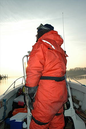 Chris Guy, wearing an orange survival suit, looks out at cold water while navigating his small boat.