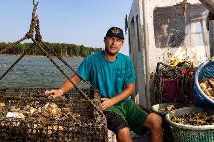 An oyster farmer in a t-shirt and cap sits between a full oyster cage and the cabin of his workboat