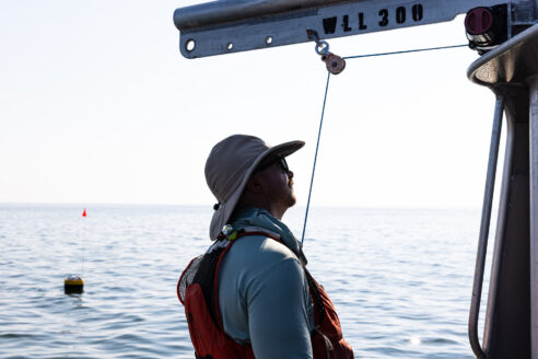 A technician in a broad-brimmed hat is silhouetted against open water, with a small buoy floating near the horizon.