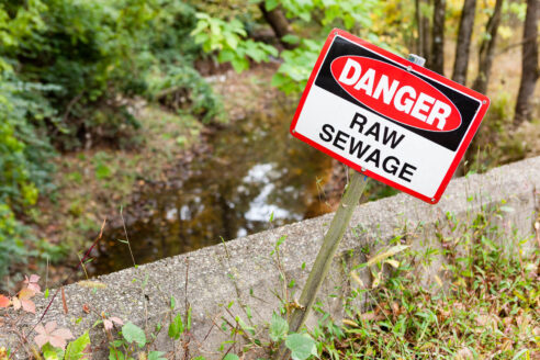 A sign reading Danger- Raw Sewage, stands in front of a creek.