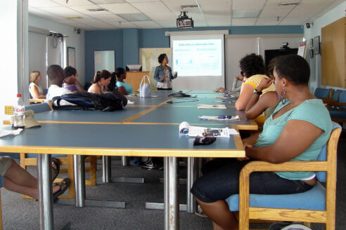 Students sit at a long conference table with a slide presentation being given.