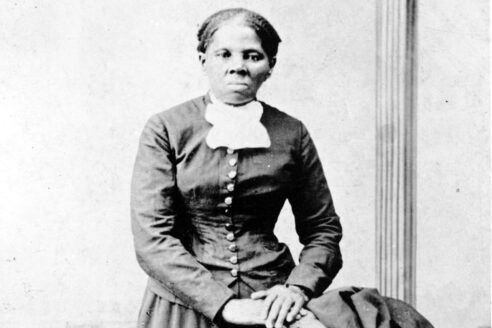 Women with hair up facing the camera. Her hands are in her lap and she is wearing a long-sleeve black ankle-length dress.