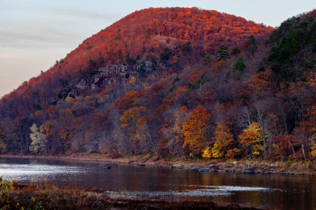 Fall foliage brightens a ridge along the Susquehanna River, where two people fish from a boat.