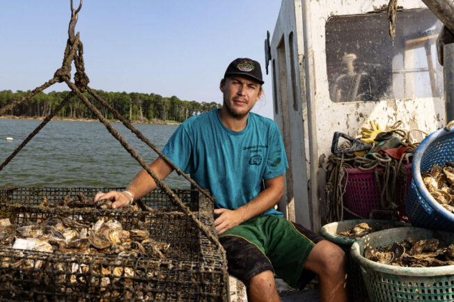 A man sits on the gunwale of a workboat, his hands resting on a cage of hatchery-raised oysters and his leg leaning against plastic bushel baskets that are full of the iconic bivalve.