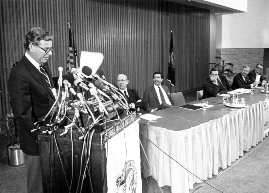 A black and white photo shows a man speaking at a podium with several news microphones attached, while other people in professional business dress listen from a long table.