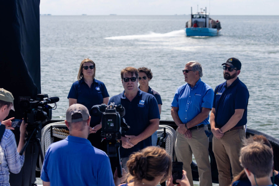 Five officials on board a boat make an announcement to a small press pool, while a large boat in the background travels out into the river.