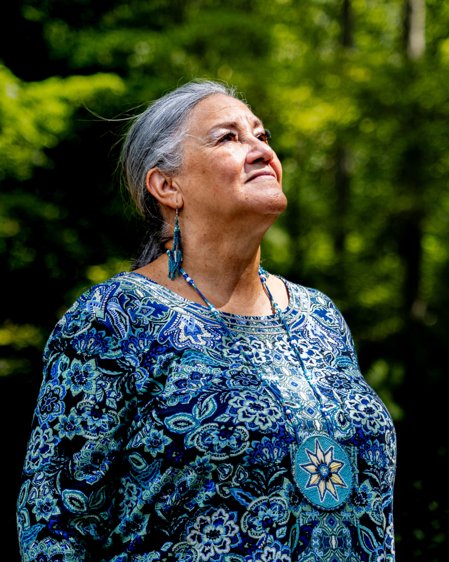 Chief Anne Richardson stands proudly, looking slightly upward, wearing a blue patterned dress with matching necklace and earrings in traditional patterns.