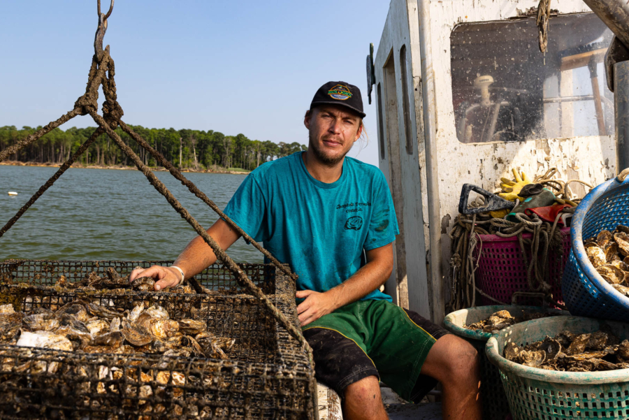 An oyster farmer sits on his dirty, cramped boat next to a cage of his oysters.