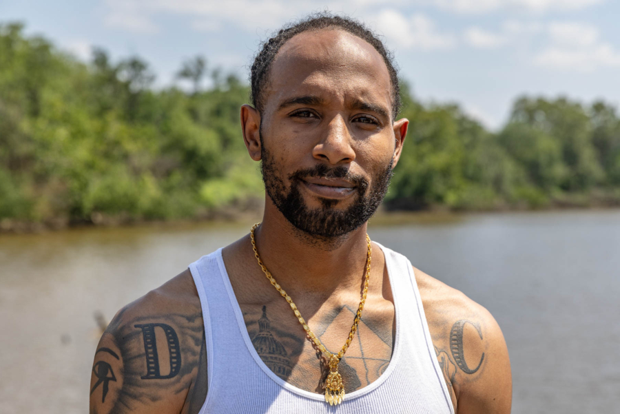 Earl Charles, with tattoos on his bare shoulders and a gold necklace over his tank top, looks warmly into the camera with the river behind him.