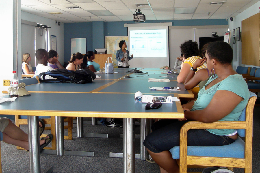 Students sit at a long conference table with a slide presentation being given.