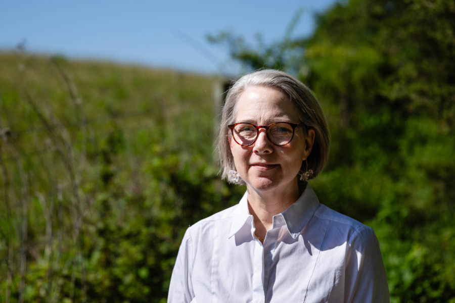 Sara, a white woman with white hair, a white shirts, and glasses, stands in front of the pasture.