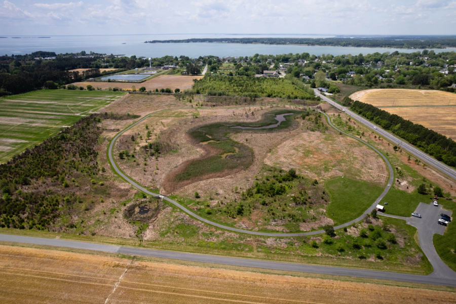 A view of a restored wetland from the air. A paved road encircles the restored wetland and water lies in the distance. Houses are scattered in-between the water and the restored wetland with trees amongst them.