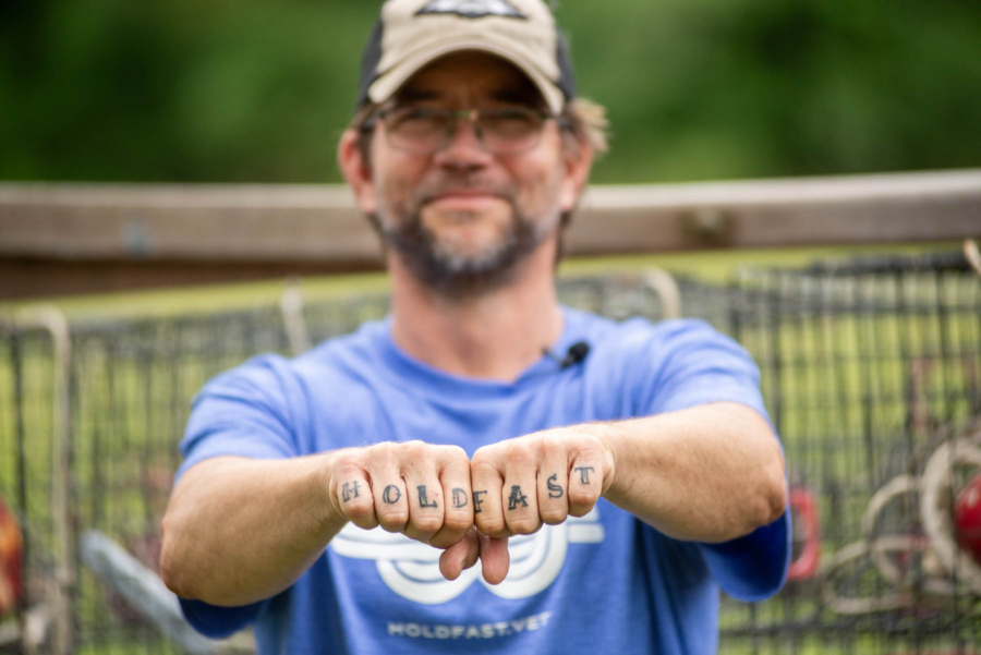 A man in a blue shirt and baseball hat displays tattoos spelling "HOLD FAST" on his knuckles.