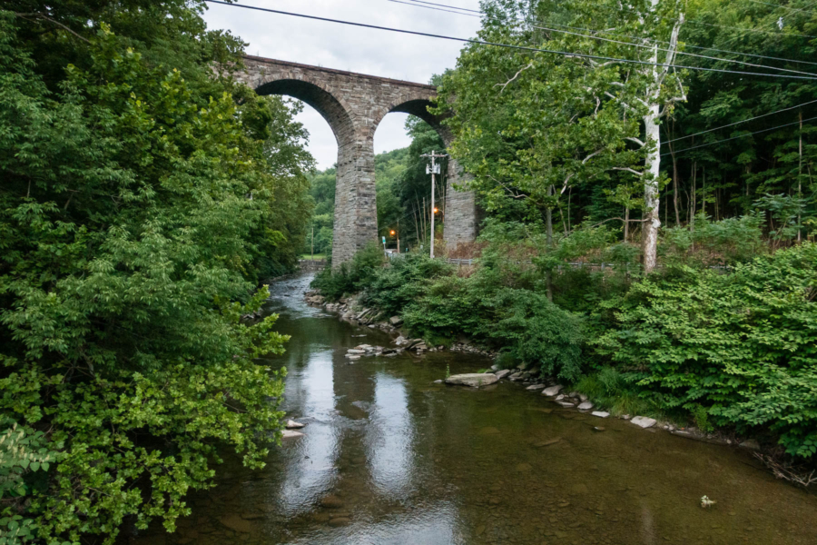 A stone railroad bridge looms high in the sky, above a stream in the forest.