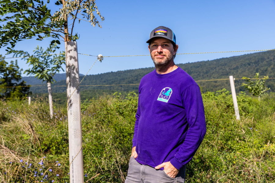 Bryan Hoffman, a white man wearing a purple shirt and blue hat, stands on a farm near recently planted trees.