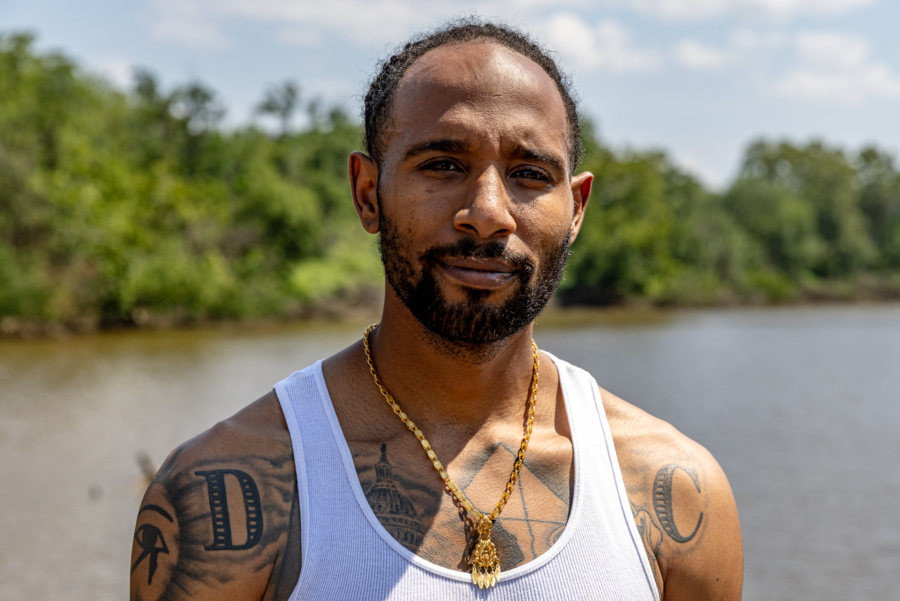 Earl Charles, a Black man approaching middle age, looks serenely into the camera with the forested bank of the Anacostia River behind him, tattoos visible around his tank top including one with an Ancient Egyptian design of an eye.
