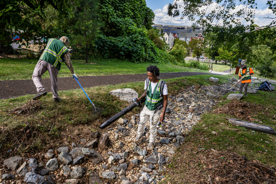 Crew members stand with rakes and leaf blowers in a rocky swale next to a pedestrian pathway, a residential neighborhood in the distance